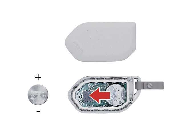 2026 Kia Tasman Smart key battery replacement image. The back cover is removed, showing the internal components. A CR2450 battery is displayed with positive (+) and negative (-) polarity indicators. A red arrow inside the battery slot points to the left, indicating the installation direction. Explains the process of replacing the smart key battery.