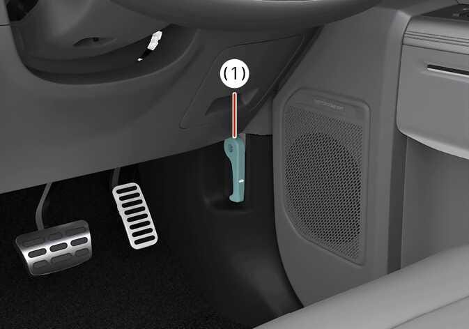 2026 Kia Tasman hood release lever image. The hood release lever (1) is highlighted in teal on the lower dashboard panel, located above the pedals and to the left of the speaker. Explains the function of releasing the hood latch from inside the vehicle.