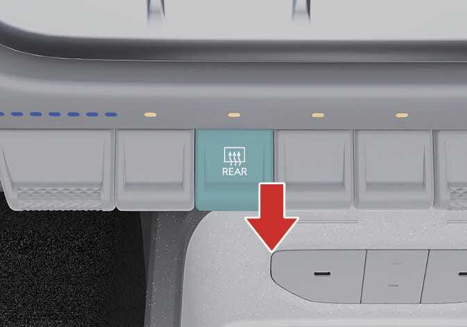 2026 Kia Tasman rear window defroster switch image. The switch labeled 'REAR' with a defrost icon is highlighted in teal among the control switches. A red arrow points downward, indicating the direction to press the switch. Explains the function of operating the rear window defroster.