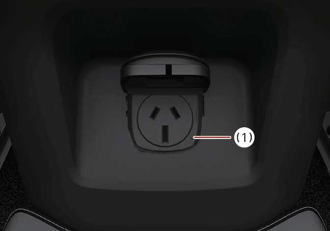 2026 Kia Tasman AC inverter outlet image. It displays the AC power socket with its cover open, located at the back of the console. A red line points to the indicator light (1) on the socket face. This explains the function where the indicator light turns on when the inverter is supplying power.