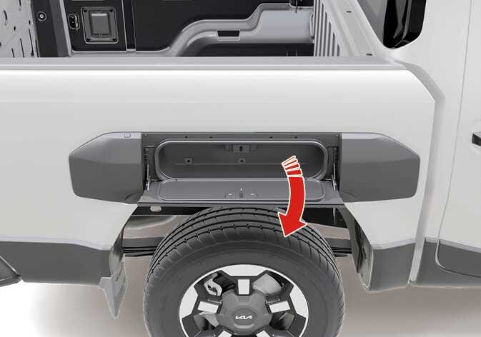 2026 Kia Tasman side storage image. The side storage compartment located above the rear wheel is shown with its door open. A red curved arrow indicates the downward opening direction. This explains the function of opening the side storage door.