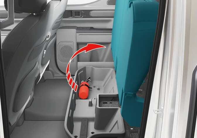 2026 Kia Tasman storage under the rear seat cushion image. The rear seat cushion, highlighted in teal, is lifted upward as indicated by a red curved arrow. A storage compartment containing a red fire extinguisher is visible underneath. Explains the function of lifting the seat cushion to access the storage space.