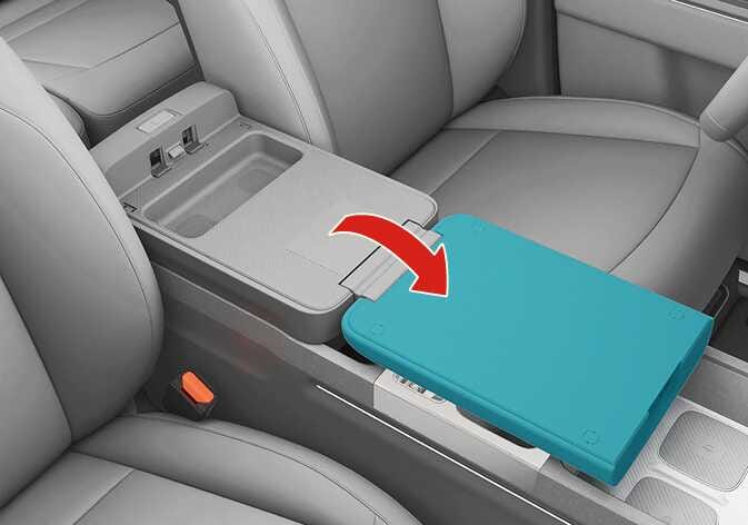 2026 Kia Tasman folding console table image. The center console armrest lid, highlighted in teal, flips forward as indicated by a red curved arrow. This illustrates the function of unfolding the armrest to use it as a flat table surface.