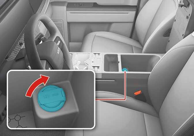 2026 Kia Tasman power outlet image. It displays the center console area between the front seats. A detailed view highlights the power outlet inside the storage in teal, with a red curved arrow indicating to lift the cover. This explains the location and operation of the power outlet.