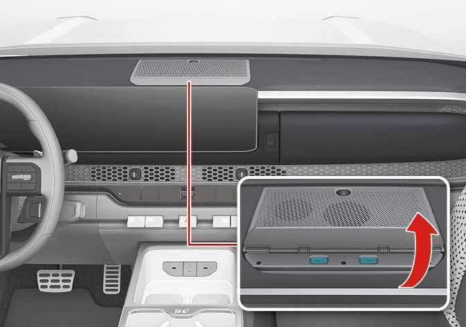 2026 Kia Tasman USB charger compartment image. A red line indicates the location of the charger on the top center of the dashboard. A detailed view shows the mesh cover lifting upwards as indicated by a red curved arrow, with cyan-highlighted release buttons on the front. Explains the function of opening the cover to access the USB charger.