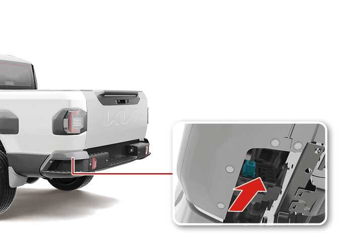 2026 Kia Tasman tailgate cable disconnection image. A close-up view of the rear bumper shows the cable connection point with a teal-highlighted connector. A thick red arrow points diagonally upward, indicating the direction to detach the cable. This explains the process of disconnecting the cable at the bottom of the rear bumper.