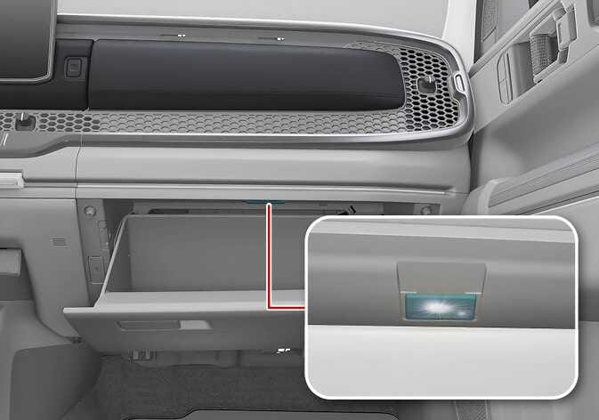 2026 Kia Tasman glove box lamp image. The passenger-side glove box is open, with a red line indicating the location of the internal light. A magnified inset shows the rectangular lamp illuminating. Describes the function where the lamp automatically turns on when the glove box is opened.