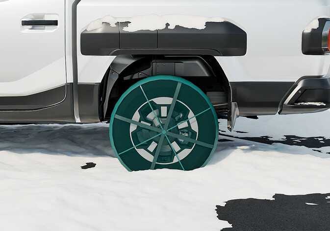 2026 Kia Tasman fabric type chain image. The rear wheel is shown on a snowy road with a fabric snow chain highlighted in teal covering the tire. The chain features a fabric cover with straps crossing the center. This illustrates the use of fabric chains for winter driving.