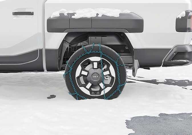 2026 Kia Tasman wire type tire chain image. A wire type snow chain, highlighted in cyan, is installed on the rear tire of the vehicle, and snow is visible on the wheel arch. Explains the appearance of the wire type chain mounted on the tire for safe driving in snowy or icy conditions.
