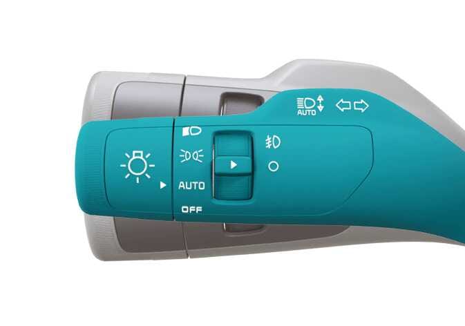 2026 Kia Tasman turn signal lever image. The lever is highlighted in teal, displaying icons for headlights, AUTO mode, fog lights, and turn signal arrows. Explains the function of operating the turn signal lever to activate the Blind-Spot View Monitor.