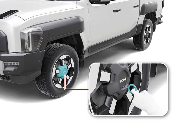 2026 Kia Tasman wheel cover removal image. The front wheel is shown with the center cap highlighted in cyan. A detailed inset shows a hand using a hook tool, highlighted in cyan, to pull the center cap outward. This describes the process of removing the wheel cover using the tool.