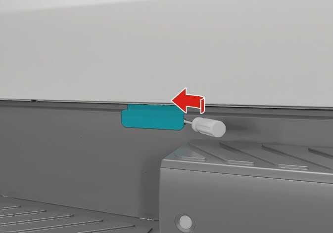 2026 Kia Tasman spare tire cover opening image. A teal-highlighted rectangular cover is on the rear bumper, with an emergency key inserted. A red arrow points left, indicating the sliding direction. Describes the function of opening the spare tire cover.