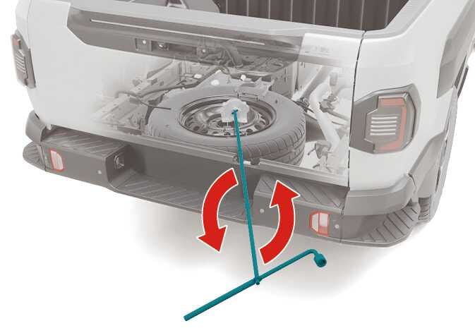 2026 Kia Tasman spare tire lowering image. A teal-colored wheel nut wrench is inserted into the access hole on the rear bumper, with red curved arrows indicating a counter-clockwise rotation. Describes the function of turning the wrench to lower the spare tire to the ground.