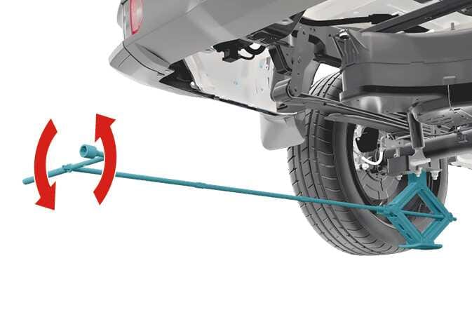 2026 Kia Tasman jack operation image. A scissor jack highlighted in cyan is positioned under the rear frame, connected to the jack handle and wheel lug nut wrench. Red curved arrows indicate the rotational direction to turn the wrench. This explains the function of operating the jack to raise the vehicle.