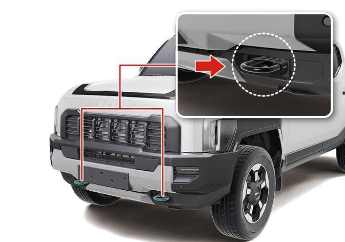 2026 Kia Tasman integrated front recovery hook image. Two hooks at the bottom of the front bumper are highlighted in teal. A magnified inset shows the hook detail within a white dotted circle, indicated by a red arrow. Describes the location of the recovery hooks for emergency off-road recovery.