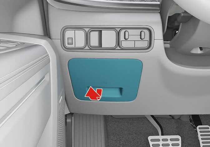 2026 Kia Tasman inner fuse panel cover image. The fuse panel cover located on the lower dashboard to the left of the steering wheel is highlighted in teal. A red arrow points outwards from the bottom handle, indicating the direction to pull. Explains the function of opening the fuse panel cover.