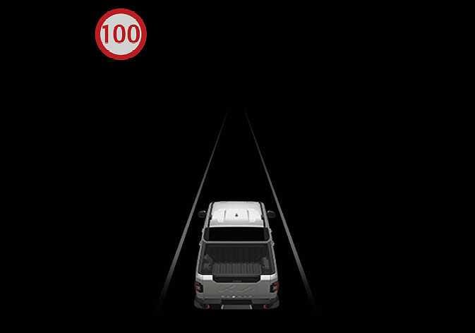 2026 Kia Tasman Intelligent Speed Limit Assist speeding warning image. A white pickup truck is shown driving on a dark road, with a speed limit sign icon displaying 100 inside a red circle in the upper left corner. This explains the function where the sign blinks and an audible warning is triggered when the vehicle exceeds the speed limit.