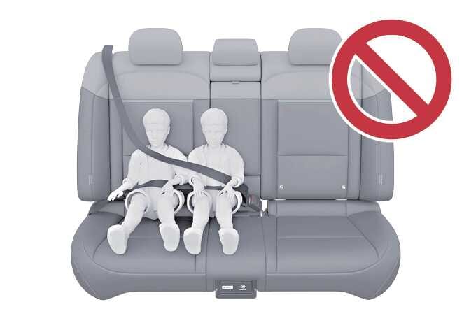 2026 기아 타스만 안전벨트 잘못된 착용 사례 이미지입니다. 뒷좌석 중앙에 두 명의 어린이가 나란히 앉아 하나의 안전벨트를 함께 착용하고 있는 모습 위에 붉은색 금지 표시 아이콘이 있습니다. 안전벨트 하나를 두 명 이상이 동시에 착용하면 위험하다는 것을 경고하는 기능을 설명합니다.