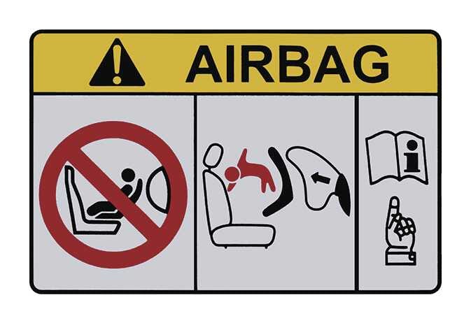 2026 기아 타스만 에어백 경고 라벨 이미지입니다. 상단에는 노란색 배경에 주의 아이콘과 AIRBAG 문구가 있습니다. 하단에는 붉은색 금지 기호로 앞좌석 어린이 보조 시트 설치 금지를 알리고, 에어백 팽창 시 붉은색으로 표시된 어린이가 충격을 받는 위험 상황을 묘사하고 있습니다. 우측에는 설명서를 참조하라는 아이콘이 함께 표시되어 어린이 탑승 시 에어백으로 인한 위험성을 경고하는 기능을 설명합니다.