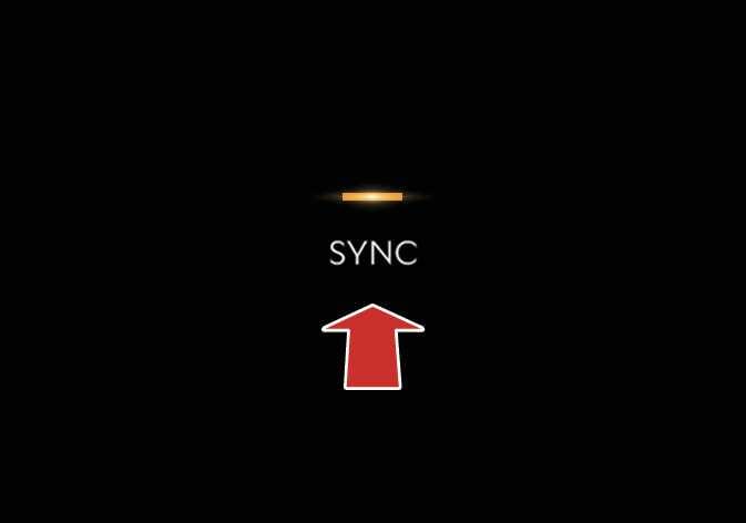 2026 기아 타스만 공조 장치 SYNC 버튼 이미지입니다. 검은색 배경에 흰색 'SYNC' 문구가 표시되어 있고, 문구 상단에는 주황색 표시등이 켜져 있습니다. 하단에는 굵은 빨간색 화살표가 위쪽을 가리키며 버튼 조작을 지시하고 있습니다. 운전석과 동승석의 온도를 동일하게 조절하기 위해 SYNC 버튼을 선택하는 기능을 설명합니다.