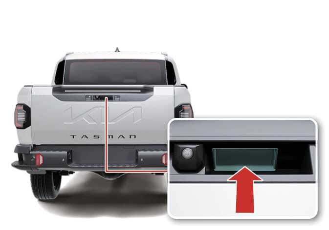 2026 기아 타스만 테일게이트 열림 버튼 위치 및 조작 방법 이미지입니다. 차량 후면 테일게이트 중앙에 위치한 버튼을 붉은색 선으로 연결하여 확대한 상세도가 포함되어 있습니다. 상세도에는 후방 카메라 우측의 열림 버튼이 청록색으로 강조되어 있으며, 하단에서 위쪽을 향하는 붉은색 화살표가 조작 위치를 지시하고 있습니다. 테일게이트 열림 버튼을 눌러 도어를 여는 기능을 설명합니다.
