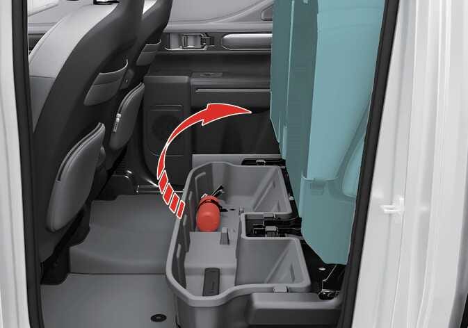 2026 기아 타스만 시트 하단 보관함 이미지입니다. 청록색으로 강조된 뒷좌석 시트 쿠션이 위로 들려 있고, 붉은색 굽은 화살표가 시트를 들어 올리는 방향을 지시합니다. 시트 아래 공간에는 소화기가 수납된 회색 보관함이 배치되어 있습니다. 시트 쿠션을 위로 올려 하단에 있는 보관함을 사용하는 기능을 설명합니다.