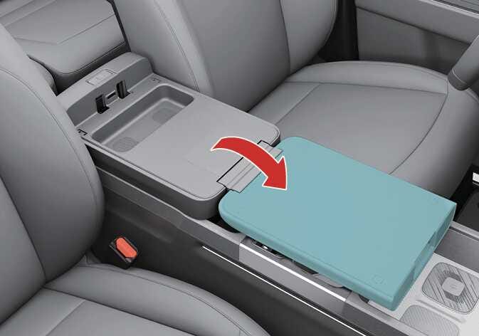 2026 기아 타스만 폴딩 콘솔 테이블 작동 이미지입니다. 센터 콘솔 암레스트 덮개가 앞쪽으로 펼쳐지는 모습이 청록색으로 강조되어 있습니다. 붉은색 굽은 화살표가 덮개를 앞쪽으로 넘기는 동작을 지시합니다. 암레스트를 열어 테이블로 사용하는 기능을 설명합니다.