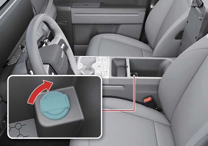 2026 기아 타스만 센터 콘솔 파워 아웃렛 이미지입니다. 운전석 옆 센터 콘솔 박스 내부를 보여주며, 하단 상세도에는 청록색으로 강조된 원형 소켓 커버가 있습니다. 빨간색 굽은 화살표가 커버를 위로 들어 올려 여는 동작을 나타냅니다. 센터 콘솔 내부에 위치한 파워 아웃렛 커버를 열어 전자기기를 연결할 수 있도록 준비하는 과정을 설명합니다.