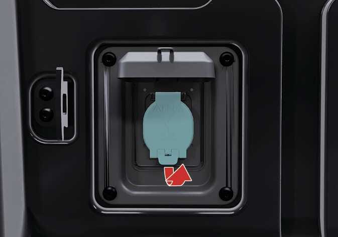 2026 기아 타스만 220 V 인버터 조작 이미지입니다. 외부 덮개가 열려 있는 상태에서 청록색으로 강조된 내부 소켓 커버 하단에 위쪽을 가리키는 붉은색 화살표가 표시되어 있습니다. 화살표 방향으로 커버를 열어 가전제품의 플러그를 연결할 수 있도록 조작하는 기능을 설명합니다.