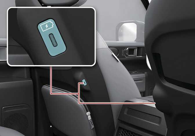 2026 기아 타스만 1열 좌석 등받이 안쪽 USB 단자 이미지입니다. 1열 시트 등받이 측면에 위치한 USB 충전 단자가 청록색으로 강조되어 있으며, 붉은색 선으로 연결된 상세도에는 번개 모양 아이콘과 USB C타입 단자 형태가 확대되어 표시되어 있습니다. 뒷좌석 탑승객이 전자기기를 충전할 수 있는 단자의 위치를 설명합니다.