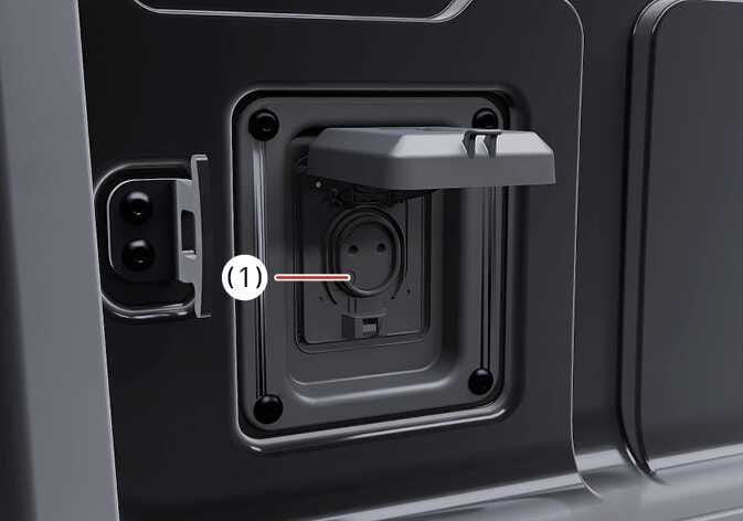 2026 기아 타스만 220 V 인버터 상세 이미지입니다. 인버터 커버가 위로 열려 있어 전원 소켓이 보이며, 소켓 옆에 위치한 작동 표시등을 번호 (1)로 지시하고 있습니다. 엔진 시동 상태에서 플러그 연결 시 표시등이 켜져 사용 중임을 알리는 기능을 설명합니다.