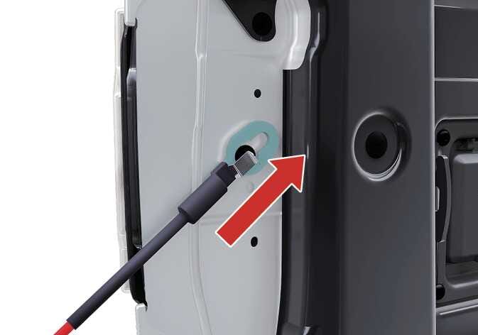 2026 기아 타스만 테일게이트 케이블 분리 이미지입니다. 차체에 고정된 테일게이트 지지 케이블의 연결 부위가 청록색으로 강조되어 있으며, 굵은 빨간색 화살표가 케이블을 위쪽 대각선 방향으로 밀어 올리도록 지시하고 있습니다. 케이블을 위로 밀어 볼트에서 분리함으로써 테일게이트를 차체에서 탈거하는 과정을 설명합니다.