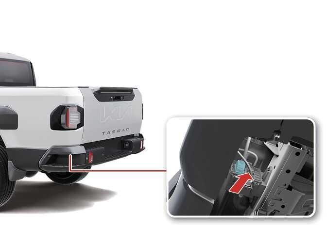 2026 기아 타스만 뒷범퍼 하단 케이블 이미지입니다. 범퍼 안쪽 상세도에 청록색으로 강조된 케이블 연결 부위가 보입니다. 붉은색 화살표가 가리키는 방향으로 조작하여 뒷범퍼 하단에 위치한 케이블을 분리하는 기능을 설명합니다.