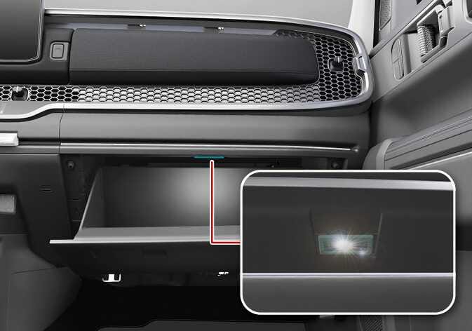 2026 기아 타스만 글로브박스 램프 이미지입니다. 조수석 대시보드 하단의 글로브박스가 열려 있고, 내부 상단에 위치한 램프가 청록색으로 강조되어 있습니다. 붉은색 선으로 연결된 우측 상세 이미지는 램프가 점등된 모습을 확대하여 보여줍니다. 글로브박스 개방 시 내부를 비추는 조명의 위치와 작동 상태를 설명합니다.