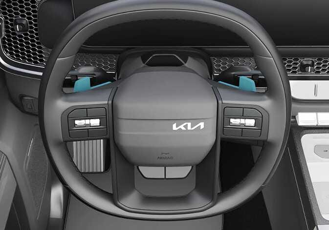 2026 기아 타스만 패들 시프트 이미지입니다. 스티어링 휠 후면 좌우측에 위치한 변속 레버가 청록색으로 강조되어 표시되어 있습니다. 스티어링 휠에서 손을 떼지 않고 기어를 변속하는 기능을 설명합니다.