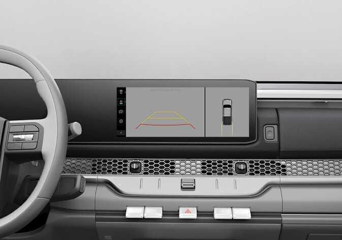 2026 기아 타스만 서라운드 뷰 모니터 작동 화면 이미지입니다. 인포테인먼트 화면 좌측에는 빨간색과 노란색 주차 가이드라인이 포함된 후방 영상이, 우측에는 차량을 위에서 내려다본 탑뷰 영상이 분할되어 표시되어 있습니다. 차량 주변 영상을 확인하여 안전하게 주차하거나 주행하도록 돕는 기능을 설명합니다.