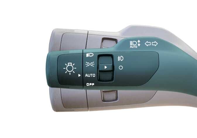 2026 기아 타스만 방향지시등 스위치 이미지입니다. 청록색으로 강조된 레버 표면에 전조등, AUTO, 안개등 조작 아이콘과 문구가 표시되어 있습니다. 레버 상단에는 상향등 조작을 의미하는 수직 화살표와 방향지시등 조작을 의미하는 좌우 화살표 아이콘이 있습니다. 방향지시등 스위치를 조작하여 후측방 모니터 영상을 켜거나 끄는 기능을 설명합니다.