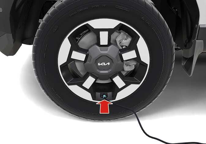 2026 기아 타스만 타이어 공기주입 밸브 연결 이미지입니다. 타이어 휠 하단의 공기주입 밸브가 청록색으로 강조되어 있고, 붉은색 화살표가 위쪽을 가리키며 호스 연결 부위를 지시합니다. 컴프레서와 타이어 연결 호스를 공기주입 밸브에 체결하는 기능을 설명합니다.