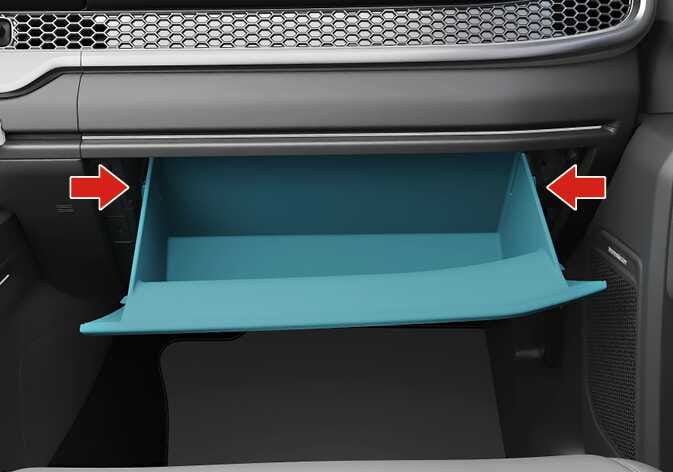 This image shows how to remove the glove box of the 2026 Kia Tasman. The glove box, highlighted in turquoise, is open, and red arrows are pointing inward on both sides. This explains how to remove the glove box by pressing inward on both sides to release the fasteners and pulling downward.