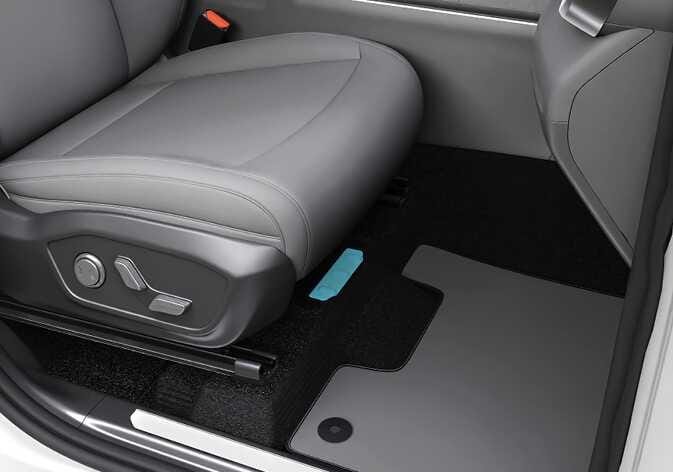 2026 기아 타스만 차대 번호(VIN) 위치 이미지입니다. 동승석 시트와 바닥면이 보이며, 시트 아래쪽 바닥 매트의 직사각형 커버 부분이 청록색으로 강조되어 있습니다. 동승석 시트 아래에 타각된 차대 번호를 확인하기 위해 바닥 커버를 찾는 위치를 설명합니다.