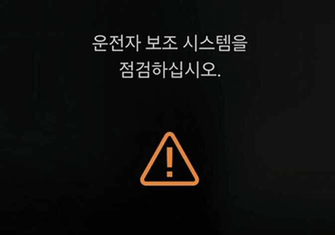 2026 기아 타스만 통합 경고 모드 화면 이미지입니다. 검은색 배경의 화면 상단에 '운전자 보조 시스템을 점검하십시오.'라는 문구가 흰색으로 표시되어 있고, 하단에는 주황색 삼각형 모양의 느낌표 아이콘이 있습니다. 차량 점검이 필요한 상황을 시각적으로 알리는 기능을 설명합니다.