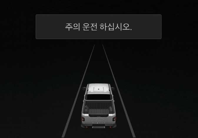 2026 기아 타스만 클러스터 경고 문구 표시 화면 이미지입니다. 어두운 배경의 화면 상단에 '주의 운전 하십시오.'라는 문구가 표시되어 있고, 하단에는 차로를 주행하는 차량의 후면 그래픽이 나타나 있습니다. 내비게이션 기반 스마트 크루즈 컨트롤 기능이 안전한 속도까지 감속할 수 없거나 진출로가 짧은 경우 등 운전자의 주의가 필요할 때 경고문이 표시되는 기능을 설명합니다.