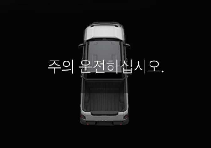 2026 기아 타스만 후방 교차 충돌방지 보조 경고 문구 이미지입니다. 검은색 배경의 클러스터 화면 중앙에 차량의 평면도가 배치되어 있고, 그 위에 '주의 운전하십시오.'라는 흰색 텍스트가 표시되어 있습니다. 긴급 제동으로 정차 시 운전자에게 주의를 당부하는 경고 문구가 표시되는 화면 구성을 설명합니다.