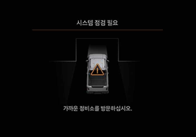 2026 기아 타스만 원격 스마트 주차 보조 기능 점검 경고 화면 이미지입니다. 인포테인먼트 화면 중앙에 차량의 평면도가 표시되어 있고, 적재함 부위에 주황색 삼각형 형태의 느낌표 경고 아이콘이 떠 있습니다. 화면 상단에는 '시스템 점검 필요', 하단에는 '가까운 정비소를 방문하십시오.'라는 안내 문구가 표시되어 있습니다. 원격 스마트 주차 보조 시스템의 이상을 시각적으로 알리고 정비소 방문을 안내하는 화면 구성을 설명합니다.