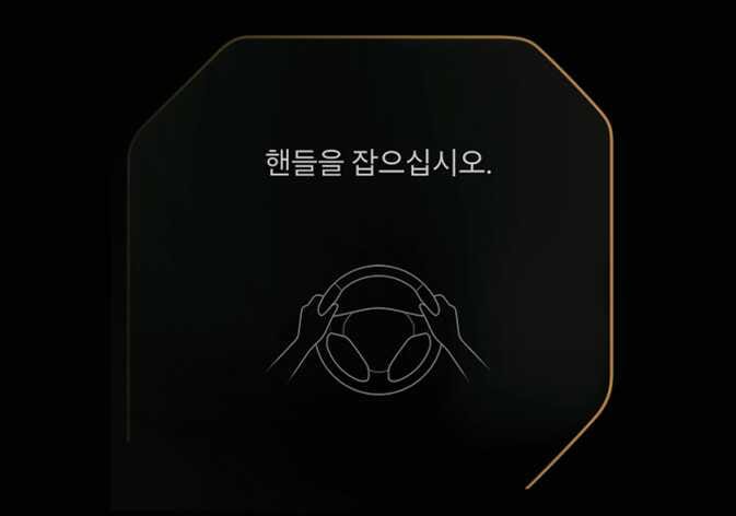 2026 기아 타스만 핸즈오프 경고 이미지입니다. 검은색 배경의 계기판 화면 중앙에 스티어링 휠을 양손으로 잡고 있는 그래픽이 표시되어 있고, 상단에 '핸들을 잡으십시오.'라는 흰색 문구가 나타나 있습니다. 일정 시간 동안 스티어링 휠을 잡지 않았을 때 운전자에게 핸들을 잡도록 경고하는 기능을 설명합니다.