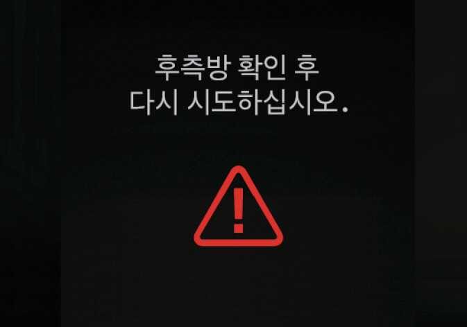 2026 기아 타스만 문 열림 방지 보조 경고문 이미지입니다. 검은색 배경의 화면 중앙에 빨간색 삼각형 모양의 느낌표 경고 아이콘이 표시되어 있습니다. 아이콘 위에는 '후측방 확인 후 다시 시도하십시오.'라는 흰색 안내 문구가 적혀 있습니다. 안전 하차 보조 기능 작동 중 후측방에서 차량이 접근할 때 문을 열려고 하면 경고하는 화면 구성을 설명합니다.