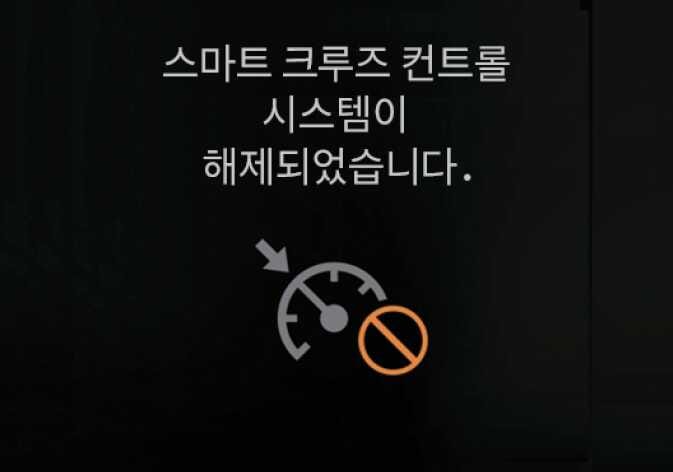 2026 기아 타스만 스마트 크루즈 컨트롤 자동 일시 해제 알림 화면 이미지입니다. 검은색 배경 화면에 '스마트 크루즈 컨트롤 시스템이 해제되었습니다.'라는 문구가 표시되어 있습니다. 하단에는 화살표가 가리키는 속도계 아이콘 위에 주황색 금지 표시가 겹쳐진 그래픽이 있습니다. 시스템이 자동으로 일시 해제된 상황을 운전자에게 경고문과 심볼로 알리는 기능을 설명합니다.