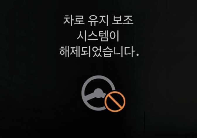 2026 기아 타스만 차로 유지 보조 해제 경고 화면 이미지입니다. '차로 유지 보조 시스템이 해제되었습니다.' 문구와 회색 스티어링 휠에 주황색 금지 표시가 겹쳐진 아이콘이 있습니다. 스티어링 휠을 잡지 않아 기능이 꺼짐을 알리는 화면 구성을 설명합니다.