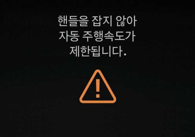 2026 기아 타스만 클러스터 경고문 이미지입니다. 화면 중앙에 '핸들을 잡지 않아 자동 주행속도가 제한됩니다.'라는 문구와 하단에 주황색 삼각형 느낌표 아이콘이 표시되어 있습니다. 운전자가 스티어링 휠을 잡지 않을 경우 주행 속도가 제한됨을 알리는 기능을 설명합니다.
