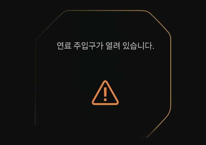 2026 기아 타스만 연료 주입구 열림 경고 이미지입니다. 검은색 클러스터 화면 중앙에 '연료 주입구가 열려 있습니다.'라는 문구가 표시되어 있고, 하단에는 주황색 삼각형 모양의 느낌표 아이콘이 위치해 있습니다. 연료 주입구가 열려 있음을 운전자에게 알리고 확인을 유도하는 기능을 설명합니다.